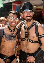 Two people smiling and wearing leather harnesses, sunglasses, and caps, standing closely together in an outdoor setting with a crowd in the background. One has tattoos and nipple piercings, suggesting a festival or parade atmosphere.