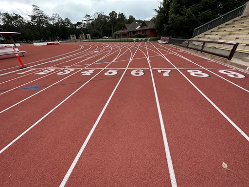 A running track with clearly marked lanes numbered from one to nine, surrounded by trees and buildings. The sky appears overcast, and there are empty bleachers to the right.