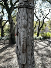 A detailed wooden carving of a bird is artfully etched into the surface of a tree or post. The carving features intricate feather details and patterns. In the background, sunlit trees with dense foliage create a natural and serene forest setting, with dappled sunlight casting shadows on the ground.
