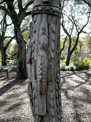 A detailed wooden carving of a bird is artfully etched into the surface of a tree or post. The carving features intricate feather details and patterns. In the background, sunlit trees with dense foliage create a natural and serene forest setting, with dappled sunlight casting shadows on the ground.