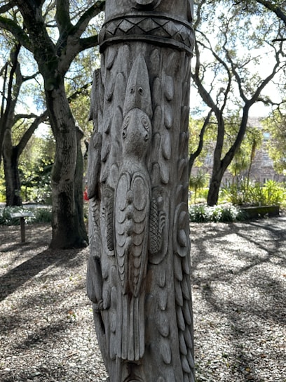 A detailed wooden carving of a bird is artfully etched into the surface of a tree or post. The carving features intricate feather details and patterns. In the background, sunlit trees with dense foliage create a natural and serene forest setting, with dappled sunlight casting shadows on the ground.