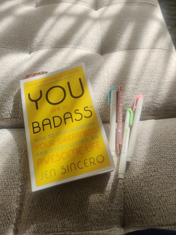 A paperback book titled 'You are a Badass' by Jen Sincero is placed on a textured cushion. Next to the book are four colored pens, positioned neatly. Sunlight casts soft shadows over the scene, giving a warm ambiance.