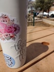 A tumbler adorned with various stickers stands on a wooden picnic table. Sunlight casts shadows across the table, with a blurred background featuring cars and trees.