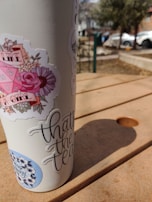 A tumbler adorned with various stickers stands on a wooden picnic table. Sunlight casts shadows across the table, with a blurred background featuring cars and trees.