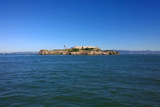A large island with a historic building sits in the middle of an expansive body of water. The island is surrounded by calm blue waters under a clear sky. The distant shoreline is visible in the background.