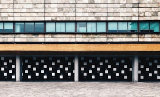 A modern architectural facade featuring a pattern of small square windows beneath a textured stone wall. Horizontal bands, including a mustard yellow strip and a series of blue-tinted glass windows, add to the geometric design. Columns support the structure, merging with a paved foreground.