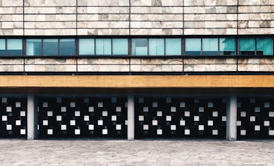 A modern architectural facade featuring a pattern of small square windows beneath a textured stone wall. Horizontal bands, including a mustard yellow strip and a series of blue-tinted glass windows, add to the geometric design. Columns support the structure, merging with a paved foreground.