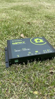 A solar charge controller branded as Newpowa MPPT is positioned on grass. The device is black with green and white text and symbols, displaying various icons related to battery and temperature. It is partially embedded in the grass, indicating an outdoor setting.