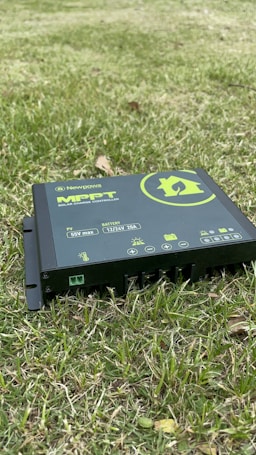 A solar charge controller branded as Newpowa MPPT is positioned on grass. The device is black with green and white text and symbols, displaying various icons related to battery and temperature. It is partially embedded in the grass, indicating an outdoor setting.