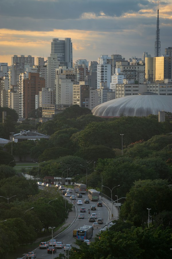 A wide São Paulo cityscape at dusk with a dense skyline of high-rise towers, a communications tower, the domed Ibirapuera gymnasium, and a tree-lined