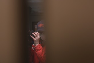 A person wearing a cap and face mask is holding a camera, partially visible through a narrow gap. The scene is dimly lit, adding a sense of intrigue or privacy.
