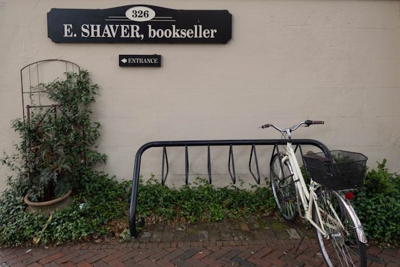 A quaint scene featuring a bicycle parked next to a black metal bike rack against a light-colored wall. Above the bicycle is a black sign with white lettering, indicating a bookseller's shop. A pot with lush green plants is placed to the left, adding a touch of greenery to the paved brick pathway in the foreground.