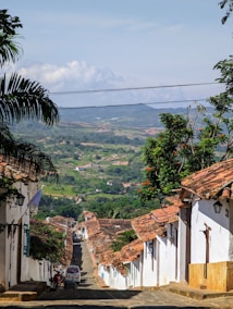 A picturesque cobblestone street lined with white colonial-style houses and terracotta roofs extends downhill. Lush greenery and rolling hills are visible in the distance under a clear blue sky. A few vehicles and a motorcycle are parked along the street, and colorful flowers and tropical foliage add vibrancy to the scene.