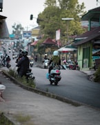 A busy street scene with several motorbikes along a winding road bordered by small shops and houses. The focus is on a few motorcyclists riding away, wearing helmets and carrying bags. The surroundings show lush greenery and urban architecture, creating an atmosphere of daily life in a bustling area.