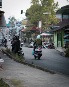 A busy street scene with several motorbikes along a winding road bordered by small shops and houses. The focus is on a few motorcyclists riding away, wearing helmets and carrying bags. The surroundings show lush greenery and urban architecture, creating an atmosphere of daily life in a bustling area.