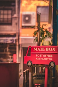 A whimsical red sign shaped like a mail delivery car is prominently displayed in the foreground. The sign reads 'MAIL BOX - POST OFFICE - MAIL DELIVERY CAR' and has a playful design with small circular details representing wheels. Behind it, an urban scene with a slightly blurred building and an air conditioning unit is visible. A small plant with green leaves is also in the background, adding a touch of nature.