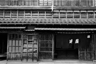 A traditional Japanese building with wooden architecture, featuring sliding doors and a fabric curtain with Japanese characters. The facade is detailed with wood panels, and a tiled roof is visible.