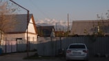 A residential area with several houses that have corrugated metal roofs. There is a parked silver car in front of a metal fence. In the background, snow-capped mountains are visible under a clear blue sky. There are some small trees and vegetation around the area.