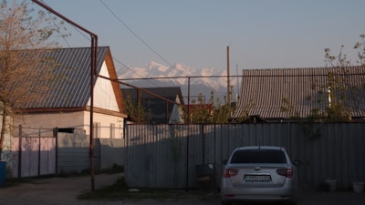 A residential area with several houses that have corrugated metal roofs. There is a parked silver car in front of a metal fence. In the background, snow-capped mountains are visible under a clear blue sky. There are some small trees and vegetation around the area.