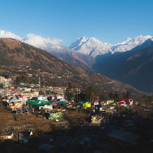 A picturesque village nestled in a mountainous region, with vibrant colored buildings scattered along the hillside. Snow-capped peaks tower in the background under a clear blue sky, and the landscape shows a mix of dense forests and terraced fields.