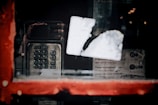 A vintage payphone with a metal keypad and screen is visible through a window with a blurred white sticker. The window has a red frame, and the reflection shows some text about costs and coin usage, suggesting a public phone booth.