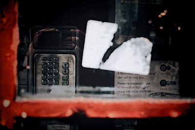 A vintage payphone with a metal keypad and screen is visible through a window with a blurred white sticker. The window has a red frame, and the reflection shows some text about costs and coin usage, suggesting a public phone booth.
