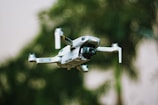 A small quadcopter drone with a camera attachment is hovering in the foreground. The background shows greenery, suggesting a natural outdoor environment with trees or plants.