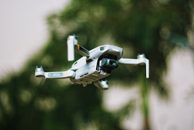 A small quadcopter drone with a camera attachment is hovering in the foreground. The background shows greenery, suggesting a natural outdoor environment with trees or plants.