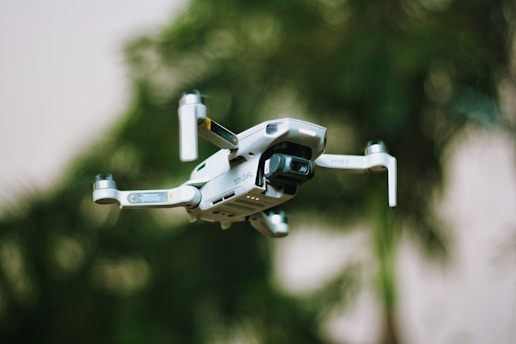 A small quadcopter drone with a camera attachment is hovering in the foreground. The background shows greenery, suggesting a natural outdoor environment with trees or plants.