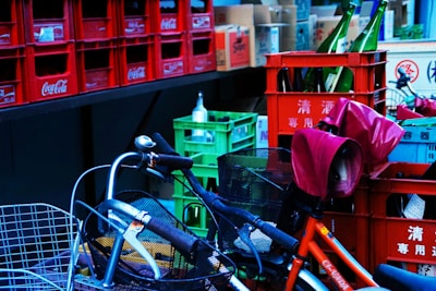 A collection of stacked crates with various bottles of different shapes and sizes. The crates predominantly bear Coca-Cola branding and are mostly red in color. Some bicycles with attached baskets and covers are parked nearby, creating a cluttered yet colorful scene.