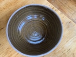 A ceramic bowl with a concentric pattern inside, featuring earthy brown and olive tones. It rests on a wooden surface with visible grain and texture.