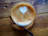 A close-up of a steaming latte art heart in a delicate ceramic cup on a polished wooden table.