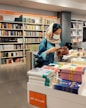 A young woman wearing a headscarf and mask is browsing a book in a bookstore. She is standing at a display shelf filled with various books, and the background is lined with bookshelves containing colorful books. The setting is bright and organized, with clear signage in an unfamiliar script.