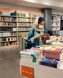 A young woman wearing a headscarf and mask is browsing a book in a bookstore. She is standing at a display shelf filled with various books, and the background is lined with bookshelves containing colorful books. The setting is bright and organized, with clear signage in an unfamiliar script.