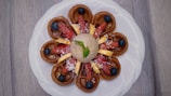 A beautifully plated dessert featuring a circular arrangement of mini waffles topped with sliced strawberries, blueberries, and banana pieces. A dollop of ice cream sits in the center with a mint leaf for garnish, all presented on a white plate against a wooden background.
