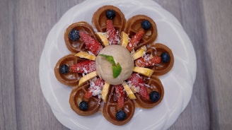 A beautifully plated dessert featuring a circular arrangement of mini waffles topped with sliced strawberries, blueberries, and banana pieces. A dollop of ice cream sits in the center with a mint leaf for garnish, all presented on a white plate against a wooden background.