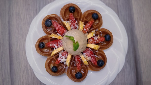 A beautifully plated dessert featuring a circular arrangement of mini waffles topped with sliced strawberries, blueberries, and banana pieces. A dollop of ice cream sits in the center with a mint leaf for garnish, all presented on a white plate against a wooden background.