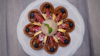 A beautifully plated dessert featuring a circular arrangement of mini waffles topped with sliced strawberries, blueberries, and banana pieces. A dollop of ice cream sits in the center with a mint leaf for garnish, all presented on a white plate against a wooden background.