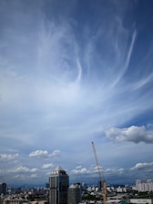 A city skyline under a vast, partly cloudy blue sky. A prominent skyscraper is towards the center, and a construction crane is visible to its right. Other buildings fill the horizon, suggesting an urban environment.