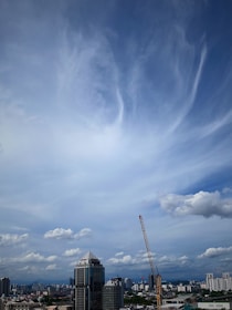 A city skyline under a vast, partly cloudy blue sky. A prominent skyscraper is towards the center, and a construction crane is visible to its right. Other buildings fill the horizon, suggesting an urban environment.