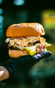 A gourmet hamburger with a glossy bun, topped with lettuce and wrapped in gold foil, held in a hand wearing a black glove, set against a blurred background.
