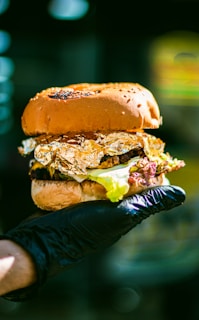A gourmet hamburger with a glossy bun, topped with lettuce and wrapped in gold foil, held in a hand wearing a black glove, set against a blurred background.