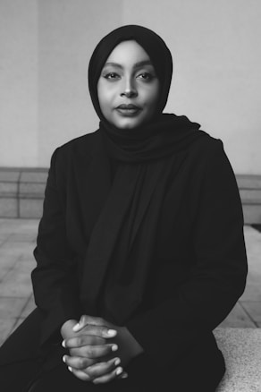 A black and white portrait of a person wearing a hijab and a dark suit, sitting with hands clasped. The background is minimal, focusing attention on the person.