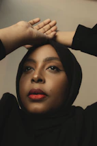 A person looks upward with hands elegantly placed on their head. They wear a black hijab and have striking red lipstick, with neutral makeup accentuating their features.