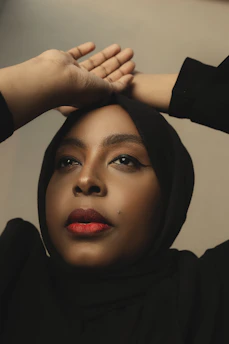 A person looks upward with hands elegantly placed on their head. They wear a black hijab and have striking red lipstick, with neutral makeup accentuating their features.
