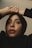 A person looks upward with hands elegantly placed on their head. They wear a black hijab and have striking red lipstick, with neutral makeup accentuating their features.