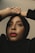 A person looks upward with hands elegantly placed on their head. They wear a black hijab and have striking red lipstick, with neutral makeup accentuating their features.