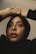 A person looks upward with hands elegantly placed on their head. They wear a black hijab and have striking red lipstick, with neutral makeup accentuating their features.