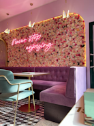 A stylish interior with a floral wall featuring a neon sign that reads 'Never stop enjoying.' The seating area includes a plush, purple tufted bench and a green chair with gold trim. The floor has a geometric patterned design, and pendant lights hang from the ceiling, adorned with butterfly decorations.