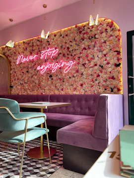 A stylish interior with a floral wall featuring a neon sign that reads 'Never stop enjoying.' The seating area includes a plush, purple tufted bench and a green chair with gold trim. The floor has a geometric patterned design, and pendant lights hang from the ceiling, adorned with butterfly decorations.